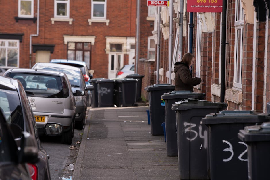 A residential street scene featuring a row of black wheelie bins lined up along the sidewalk adjacent to a red-brick building with multiple small white-framed sash windows. The bins have closed lids a